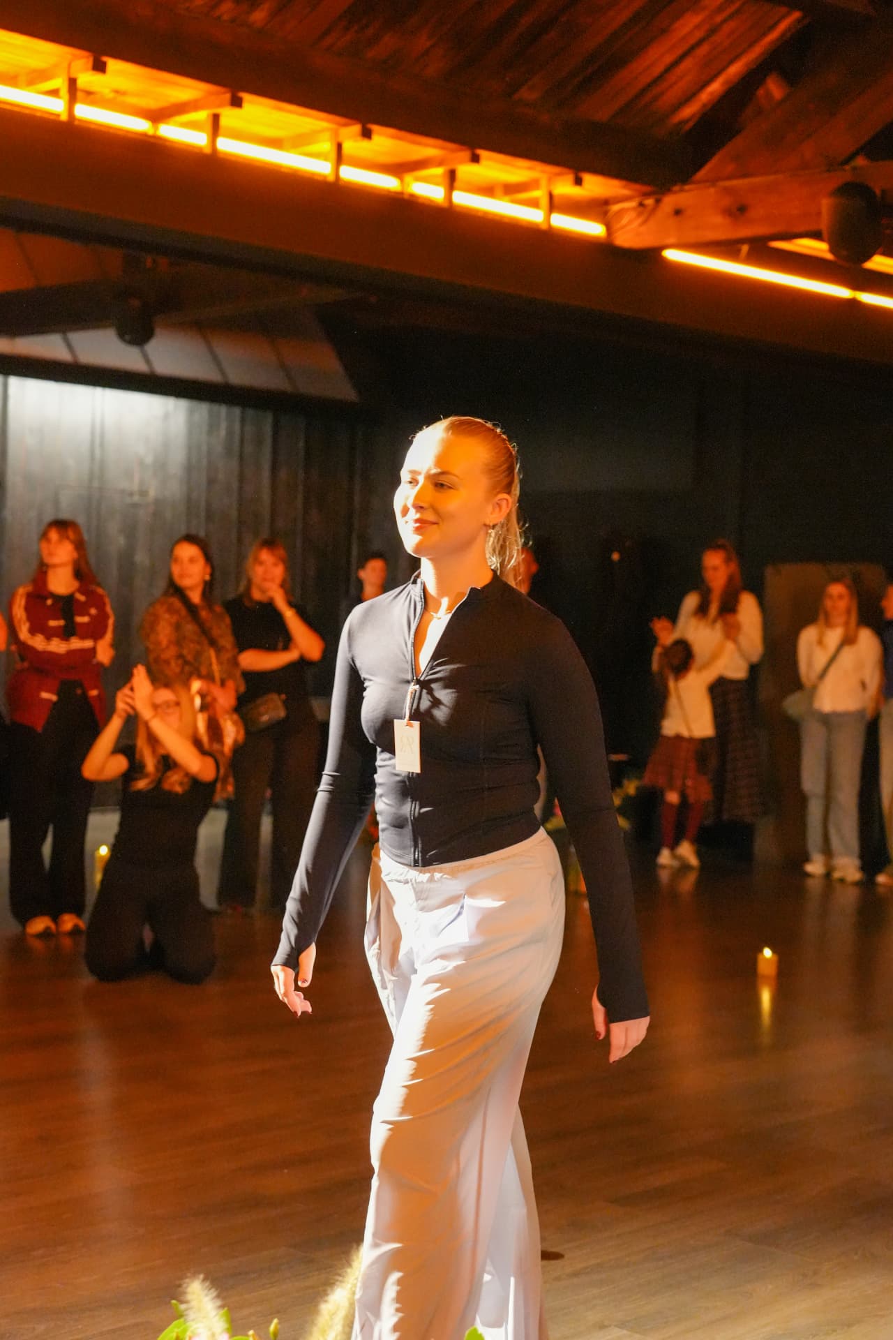 Een model in een zwart sportjasje en wijde witte broek wandelt langs het publiek