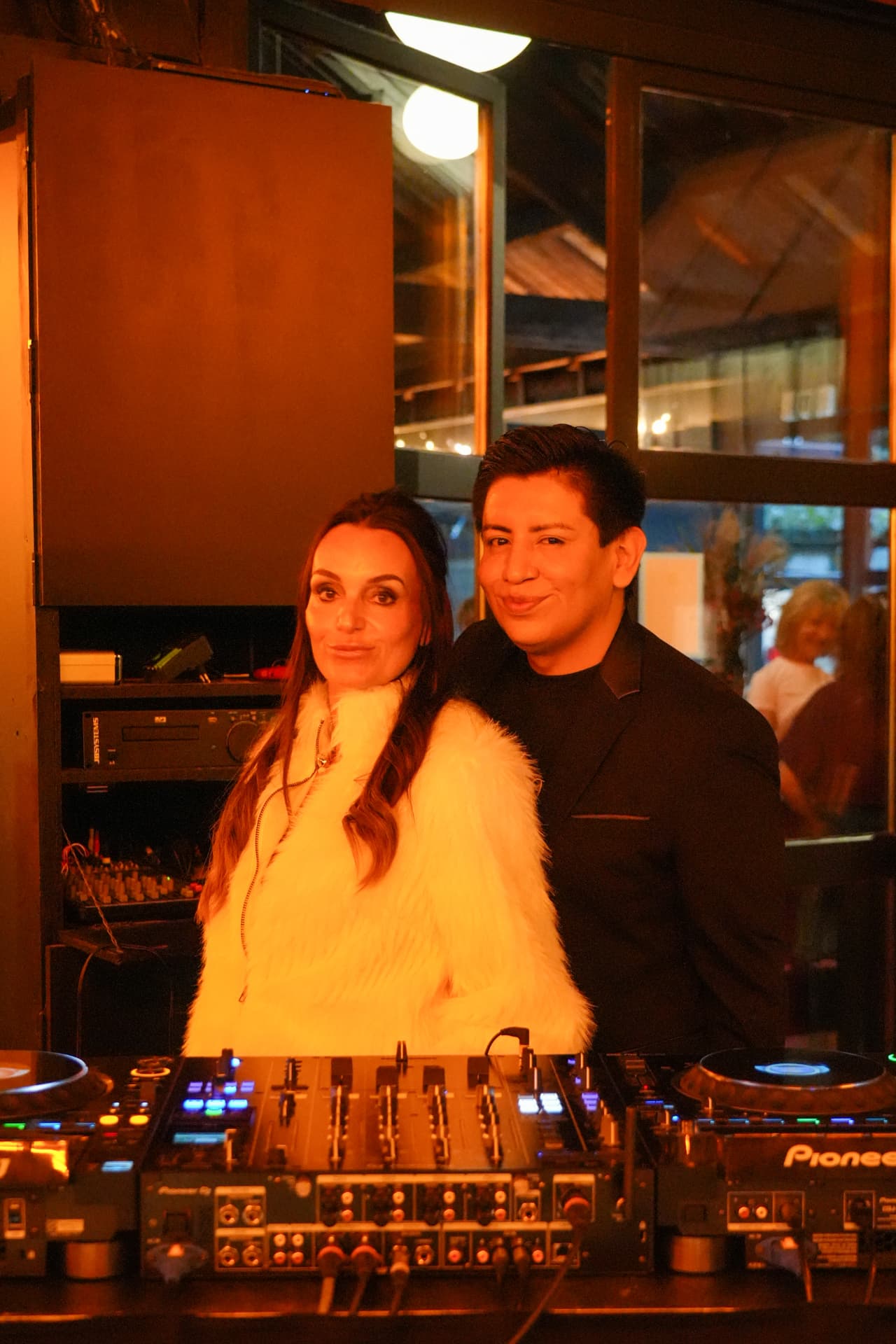 Een bezoekster in een witte jas poseert met de dj achter het mengpaneel