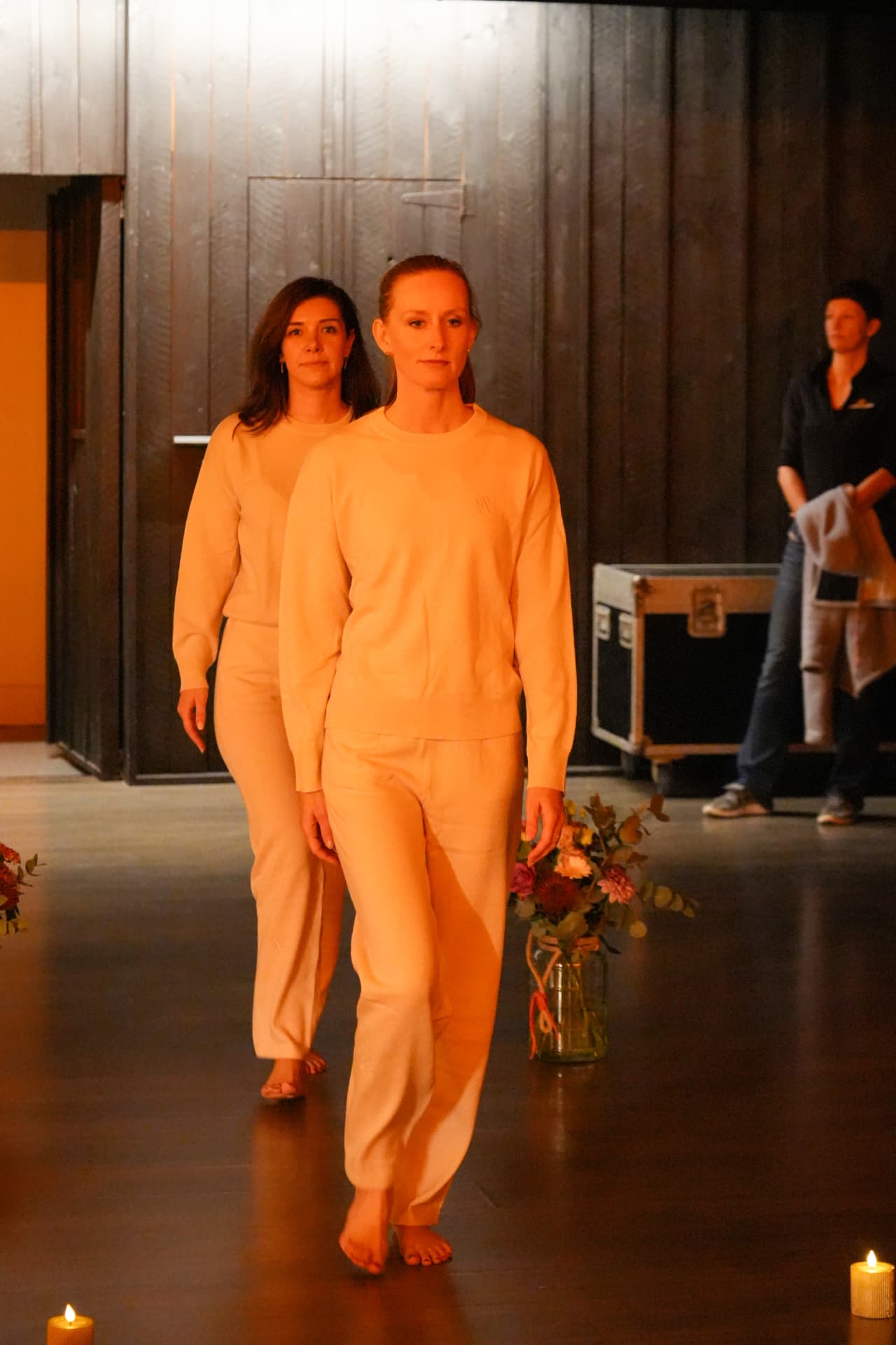 Twee modellen wandelen achter elkaar in lichte sweaters en broeken